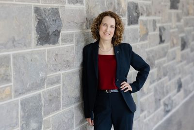 Amy Sebring Amy Sebring, wearing a suit, leans against a stone wall and smiles with a hand on her hip.